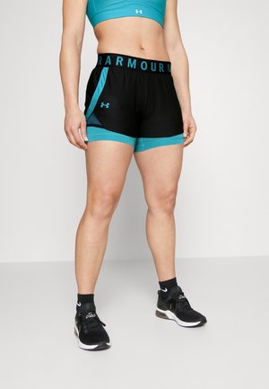 Sportowiec noszący czarne i turkusowe spodenki Under Armour, turkusowy biustonosz sportowy, czarne skarpetki i czarne buty do biegania, stojący na jednolitym tle.