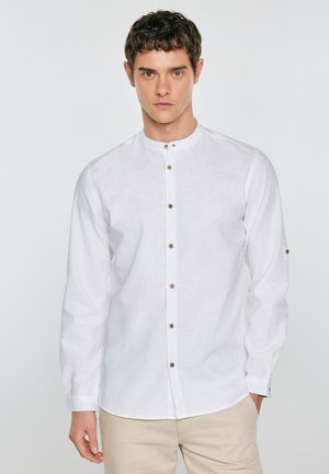 Młody mężczyzna w białej koszuli z kołnierzem mandaryńskim i beżowych spodniach, stojący na tle jednolitego jasnoszarego tła.