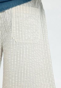 Shorts en tissu rayé de couleur claire présentant une poche à rabat, une texture structurée et des lignes verticales dans des nuances de blanc et de gris.