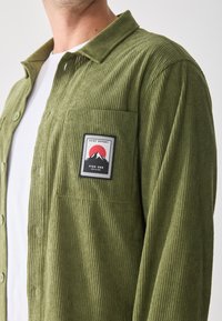 Green corduroy shirt with a buttoned front, collar, and a chest pocket featuring a rectangular patch with mountains and text.