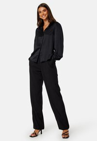 Blusa nera in satin con maniche lunghe e svasate, chiusura con bottoni sul davanti e colletto rotondo, abbinata a pantaloni neri a vita alta e scarpe con cinturini.