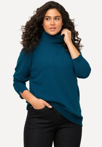 Mėlynas turtleneko megztinis su raukšlėmis, nusileidusiomis petnešomis ir šoninėmis plyšėmis, derintas su tamsiomis džinsinėmis kelnėmis. Lygus megztas audinys.