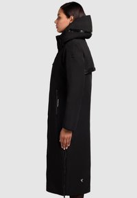 Black long coat with a high collar and oversized hood. Features zipper pockets and a textured fabric with a matte finish.