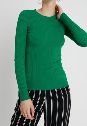 Vrouw in een aansluitend groene geribbelde top met lange mouwen en zwarte broek met verticale witte strepen, poseert met één hand bij haar nek.
