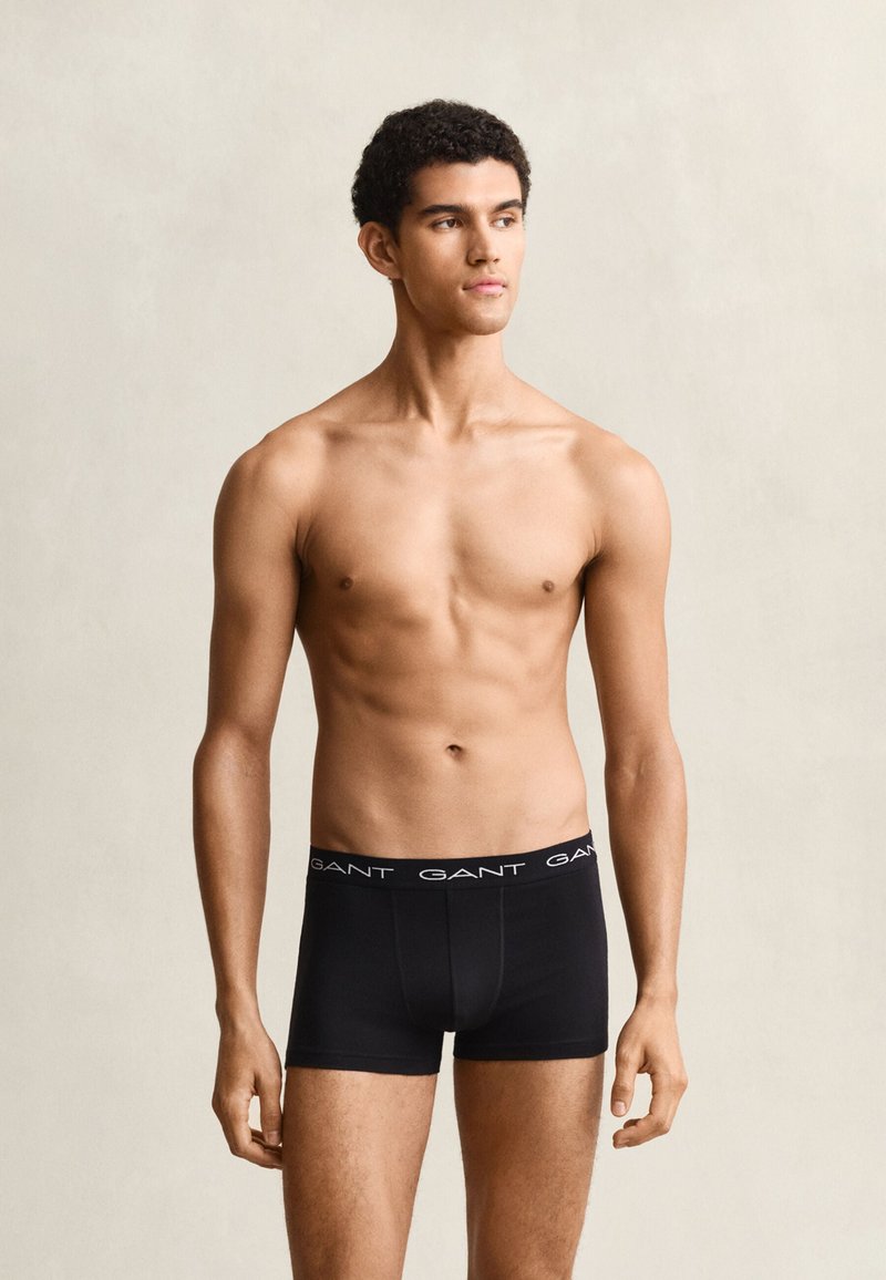 Jeune homme debout sur un fond uni portant un boxer noir GANT, regardant sur le côté avec une expression neutre.