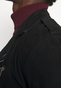 Veste noire avec une finition texturée et une fermeture éclair robuste, comportant un col roulé côtelé bordeaux en dessous. Accents de matériel visible sur les épaules.