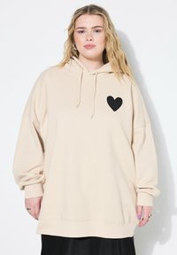 Sudadera beige de gran tamaño con capucha ajustable y estampado de corazón negro en el lado izquierdo. Tejido suave con puños y dobladillo de canalé.