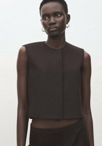 Zwarte cropped top zonder mouwen met een gestructureerd ontwerp en een voorste naad. Model draagt gouden oorringen. Gestructureerde achtergrond.