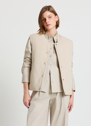 Beige leichte Jacke mit rundem Ausschnitt, Druckknöpfen und langen Ärmeln. Getragen über einem passenden, geknöpften Hemd und weit geschnittenen Hosen.
