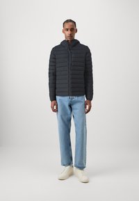 Zwarte pufferjack met horizontale stiksels, ritssluiting en een capuchon, gecombineerd met lichtblauwe rechte jeans en witte sneakers.