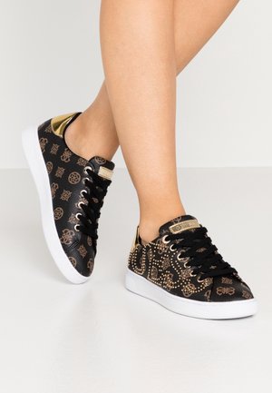 Ben iført sorte lave sneakers med brune mønstre, gyldne accenter, gyldne nitter på siderne, sorte snørebånd og hvide såler på et lyst gulv.