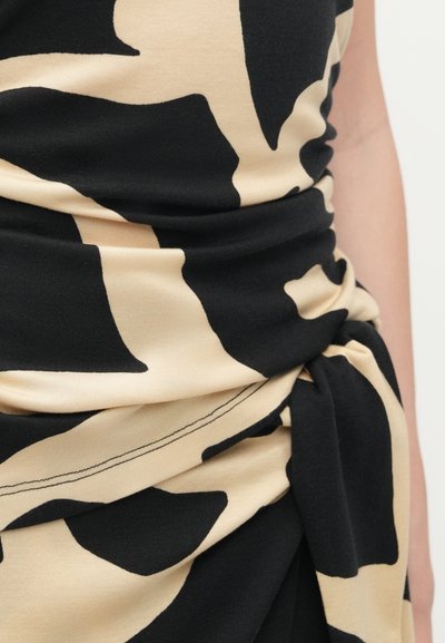 Primo piano di un tessuto a motivi astratti nero e beige raccolto e annodato in vita, che avvolge il lato e il braccio di una persona.