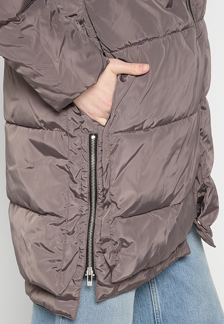 Hand in die Tasche einer glänzenden, taupefarbenen Steppjacke mit sichtbarem silbernen Reißverschluss, die über einer hellblauen Jeanshose getragen wird.