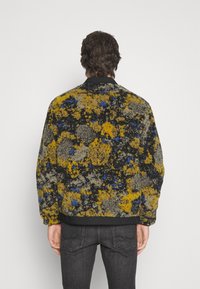 Teksturowana kurtka bomberka z wielokolorowym wzorem w odcieniach żółci, czerni i niebieskiego, z czarnym prążkowanym dołem i kołnierzem.