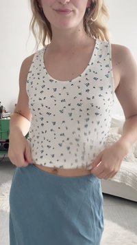 Femme portant un haut blanc sans manches à motifs floraux et une jupe bleue, ajustant ses vêtements dans une pièce lumineuse avec un lit et des meubles verts.