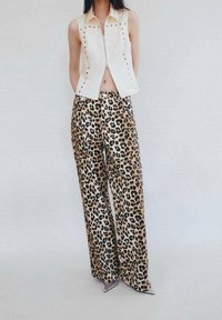 Weißer Reißverschluss-Westen ohne Ärmel mit Nietenverzierungen, kombiniert mit weit geschnittenen Leopard-Print-Hosen in Beige und Schwarz. Glatte Textur insgesamt.