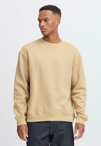 Beige crewneck sweatshirt with a relaxed fit, ribbed cuffs, and hem. Soft fabric with a smooth texture. Paired with dark jeans.