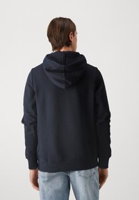 Uomo in piedi con le spalle rivolte, indossa una felpa nera con cappuccio e jeans di denim blu chiaro, contro uno sfondo neutro.