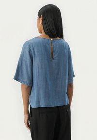 Denim bluzica s kratkimi rokavi, okroglim izrezom in detajlom z ključavnico na hrbtni strani, ki se zapre z gumbom. Mehka modra tkanina z gladko teksturo.