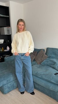 Γυναίκα που στέκεται σε εσωτερικό χώρο φορώντας ένα πλεκτό εκρού πουλόβερ, φαρδιά μπλε τζιν παντελόνια και μαύρα παπούτσια δίπλα σε ένα μπλε σαλονάκι από κορδόνι με καφέ μαξιλάρια.