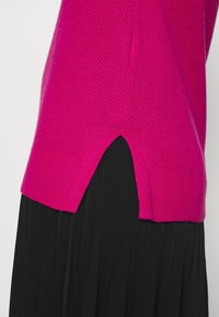 Jasnoróżowy sweter dziany z bocznymi rozcięciami noszony na czarnej plisowanej spódnicy, ukazujący tekstury materiału i warstwy odzieży.