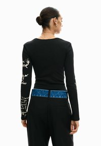 Zwart shirt met lange mouwen voorzien van witte handgeschreven tekst op beide mouwen, gecombineerd met zwarte broek met een tailleband met blauwe denimaccenten.