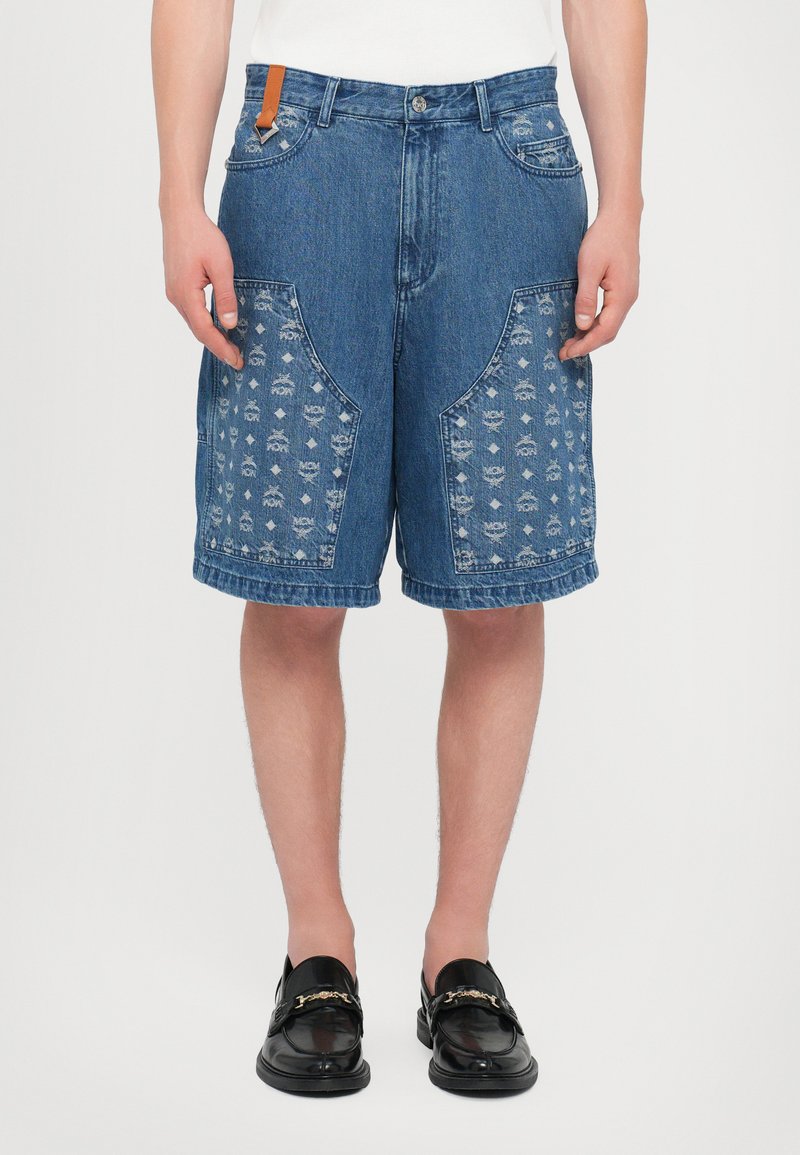 Denim shorts with large patterned pockets, blue fabric, decorative stitching, and a tan leather tag at the waistband; knee-length fit.