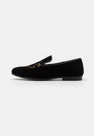 Mocassin noir en velours facile à enfiler avec un talon bas et un motif décoratif brodé en or sur le dessus avant.