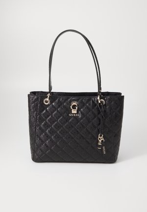 Black quilted Guess tote bag with double handles, gold logo plaque, and a hanging keychain charm on a plain background.