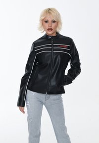 Giacca nera in ecopelle con tre strisce bianche sul petto, colletto alzato e logo rosso ricamato. Indossata con jeans azzurri chiari strappati.