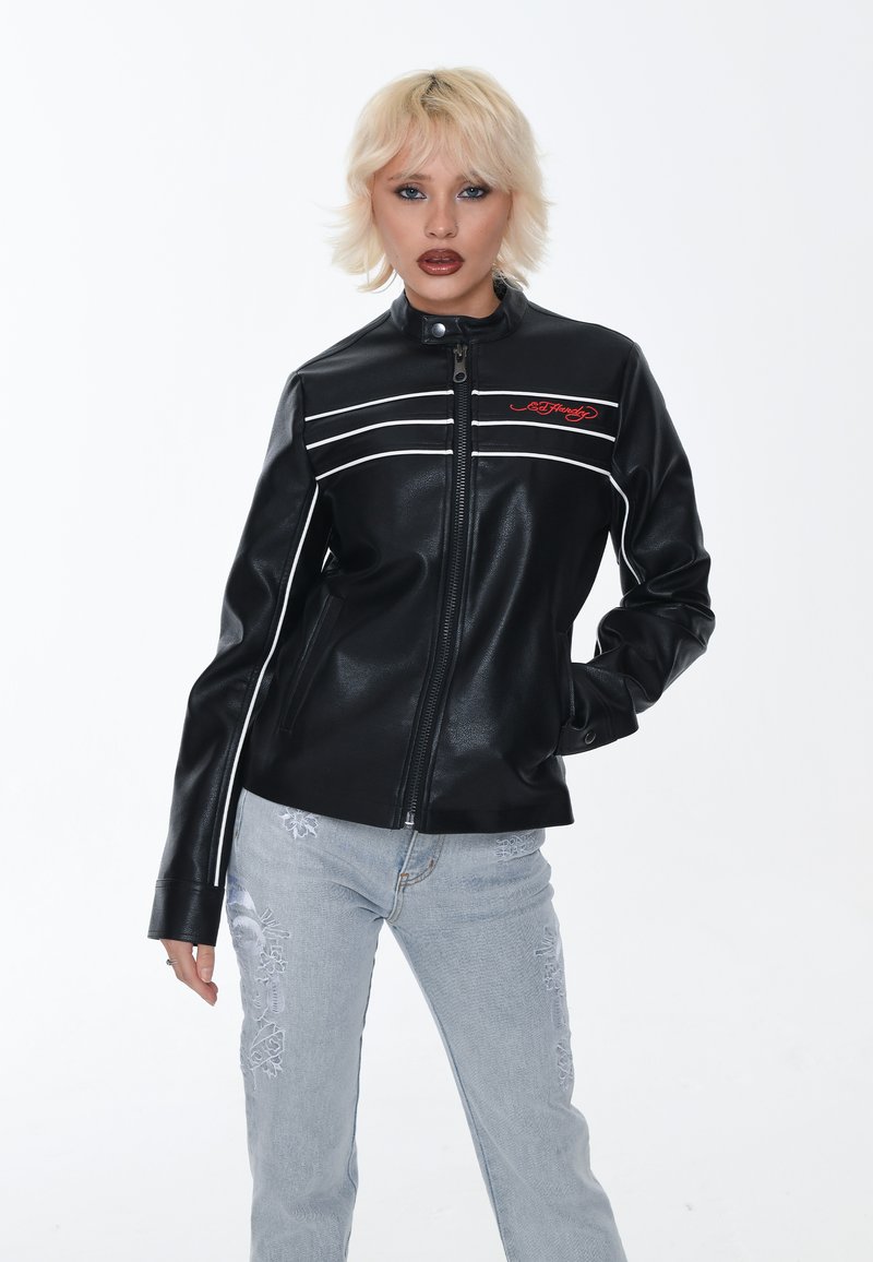 Giacca nera in ecopelle con tre strisce bianche sul petto, colletto alzato e logo rosso ricamato. Indossata con jeans azzurri chiari strappati.