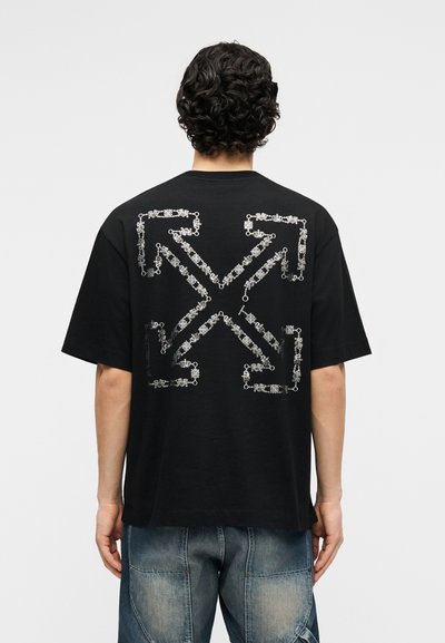 Άνδρας με σκούρα σγουρά μαλλιά που φοράει μαύρο μπλουζάκι με λευκό σχέδιο βέλους από μικρές αλυσίδες στην πλάτη, σε συνδυασμό με μπλε τζιν.