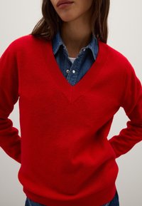 Maglione rosso in lana con scollo a V e finiture a costina, abbinato a una camicia di jeans. Il maglione presenta una texture morbida e una vestibilità rilassata, enfatizzando il concetto di sovrapposizione casual.