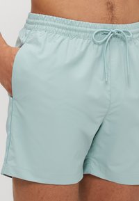 Shorts de bain bleu clair fabriqués en tissu lisse et léger. Dotés d'une taille élastique avec cordon de serrage et de poches latérales.