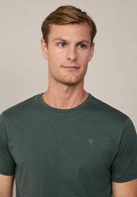Tmavo zelené tričko s krátkym rukávom, guľatým výstrihom a jemne vyšitým logom na ľavej hrudi. Mäkka a hladká textúra tkaniny.