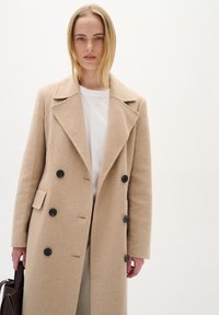 Sieviete, kas valkā beļģu krāsas divu rindu vilnas mēteli pār baltu kreklu, tur tumši brūnu rokassomu pret vienkāršu gaišu fonu.