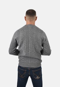 Maglione grigio lavorato a maglia con motivo a trecce, scollatura rotonda e orlo a coste, abbinato a jeans blu scuro con dettagli di cucitura visibili.