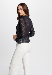Top in pizzo blu navy con maniche lunghe, dettagli floreali intricati e orlo scalloppato. Abbinato a pantaloni bianchi a gamba larga.