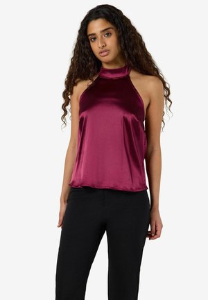 Femme aux longs cheveux bouclés portant un haut dos nu brillant rose foncé et un pantalon noir, debout devant un fond uni.
