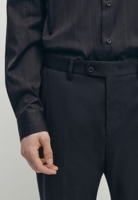 La camicia scura color navy presenta righe verticali, con maniche lunghe e polsini abbottonati. Pantaloni sartoriali con frontale piatto e chiusura a bottone.
