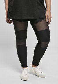 Leggings noirs avec texture côtelée et panneaux en maille transparente. Associés à des baskets blanches. La tenue comprend un long haut noir.