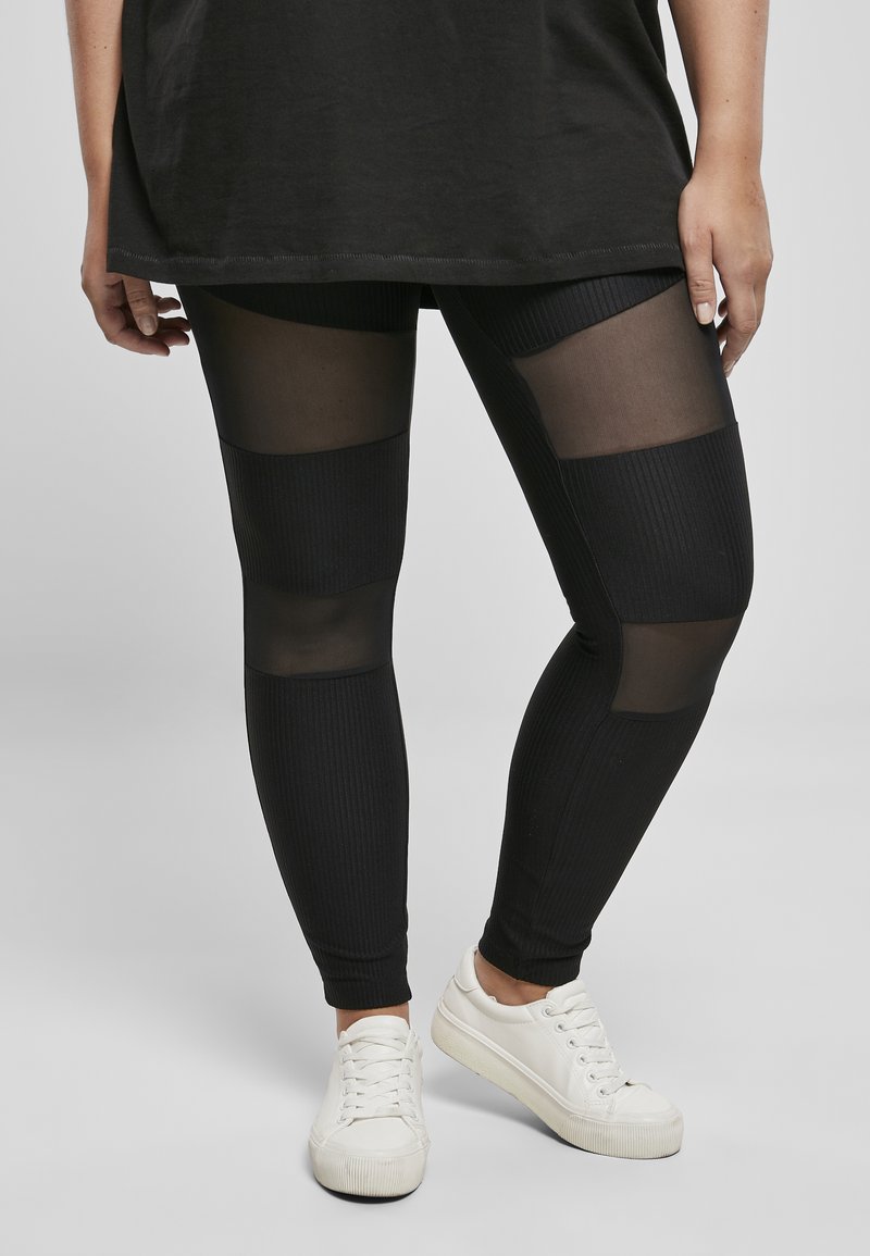 Leggings noirs avec texture côtelée et panneaux en maille transparente. Associés à des baskets blanches. La tenue comprend un long haut noir.