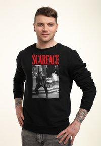 Schwarzer Sweatshirt mit einem frontal aufgedruckten "Scarface"-Grafik in rotem Text und einem schwarz-weißen Bild eines Charakters mit einer Waffe.