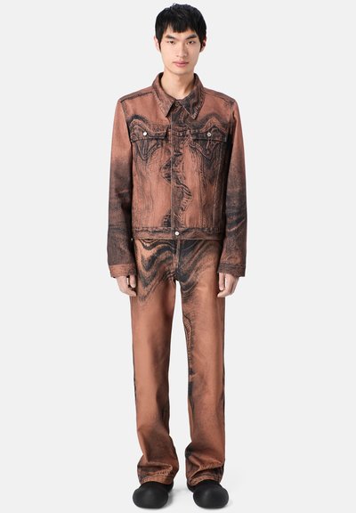 Man som bär en matchande brun och svart acid-wash denimjacka och byxor, står med avslappnade armar, svarta slip-on skor, neutral bakgrund.
