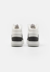 Tenisky s vysokým svrškem z bílé textilie, černými textilními panely a texturovanou podrážkou. Mají polstrovaný límec a minimální množství značkových prvků.