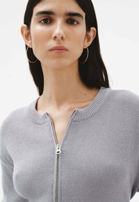 Suéter gris de punto con cuello redondo y cremallera frontal completa. Presenta un cuello y puños de canalé, textura suave y corte ajustado.