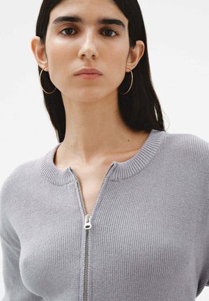 Suéter gris de punto con cuello redondo y cremallera frontal completa. Presenta un cuello y puños de canalé, textura suave y corte ajustado.