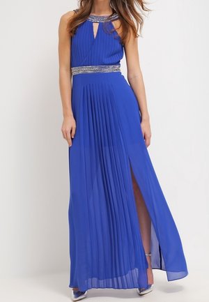Mouwloze koningsblauwe geplooide jurk met een met juwelen versierde halslijn en tailleband, vooraan een split die zilveren hakken met enkelbandje onthult.