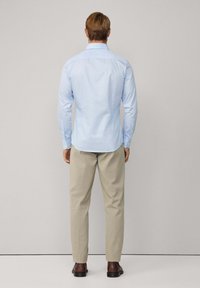 Man staat met zijn rug naar ons gekeerd, draagt een lichtblauw overhemd, beige broek en donkerbruine schoenen voor een effen lichtgrijze achtergrond.