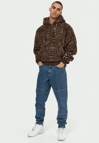 Jovem vestido com um hoodie de fleece castanho com padrões, jeans em remendos azuis e tênis brancos, de pé com as mãos nos bolsos em fundo branco.